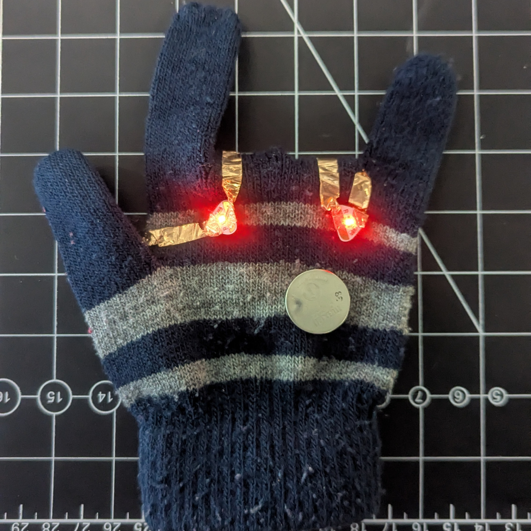 Here is the wearable design from the front view, with the battery on the other side of the gloves. The reason there's another battery is that when I tried to take a picture, the other one wouldn't light up unless I pushed it down, so I just used my extra battery to keep the LED working.